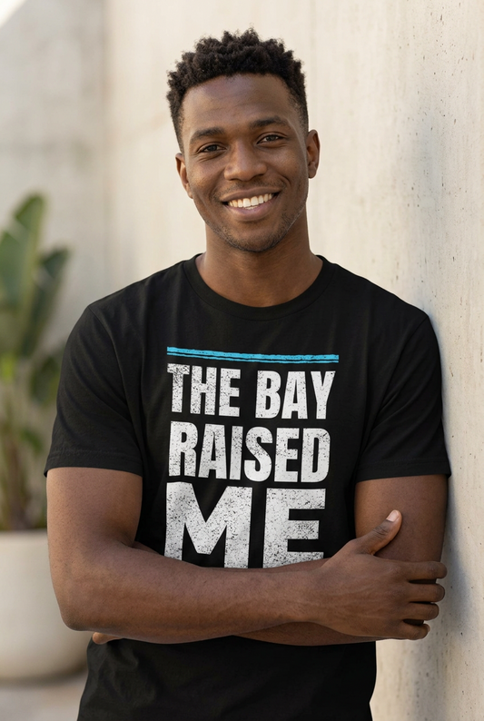 A smiling man wearing a black graphic t-shirt with the text 'THE BAY RAISED ME' in distressed white letters and a thin blue horizontal line above, standing against a light-colored wall.