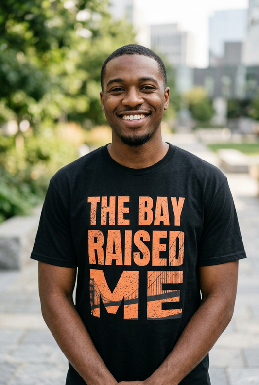 A smiling man wearing a black t-shirt with a distressed orange graphic that reads 'THE BAY RAISED ME' in an outdoor setting.