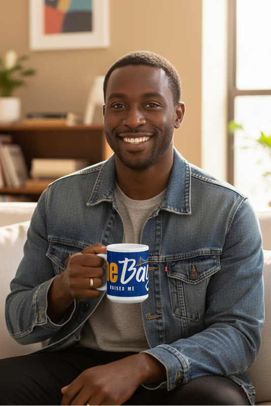 A smiling man in a denim jacket sits on a couch holding a blue and white ceramic mug with the text 'Be Baptized Raised Me' in a living room setting.